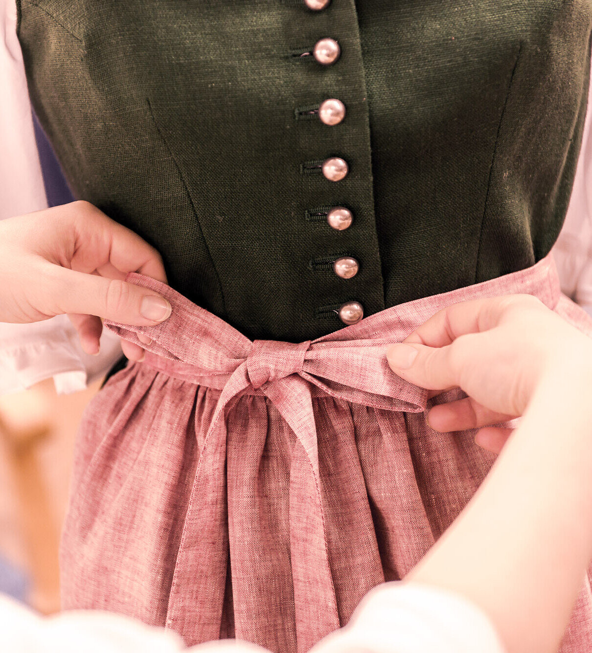 Hände binden die Schleife einer rosafarbenen Schürze an einem grünen Dirndl mit weißer Bluse – Detail traditioneller Tracht.