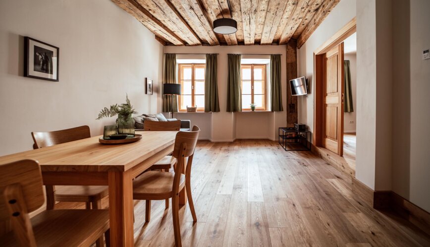 Heller Wohn- und Essbereich mit rustikaler Holzdecke, Parkett und Holztisch; grüne Vorhänge, Sofa und Wand-TV sorgen für gemütliches Ambiente.