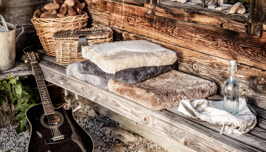 Gestapelte Lammfell-Sitzkissen in Naturtönen auf einer Holzbank vor der Hütte, daneben Weidenkörbe mit Zapfen, eine Gitarre und eine Bügelflasche.