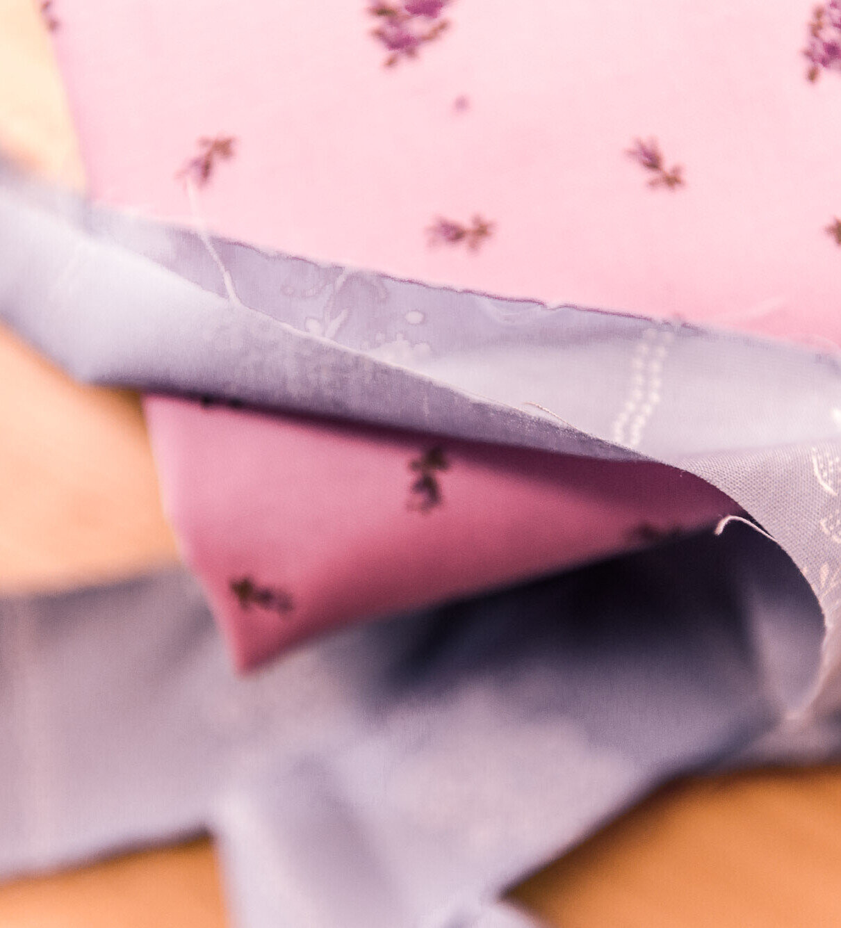 Rosa Stoff mit kleinen lila Blüten, dazu ein graues Band mit feiner Stickerei auf hellem Holzhintergrund.