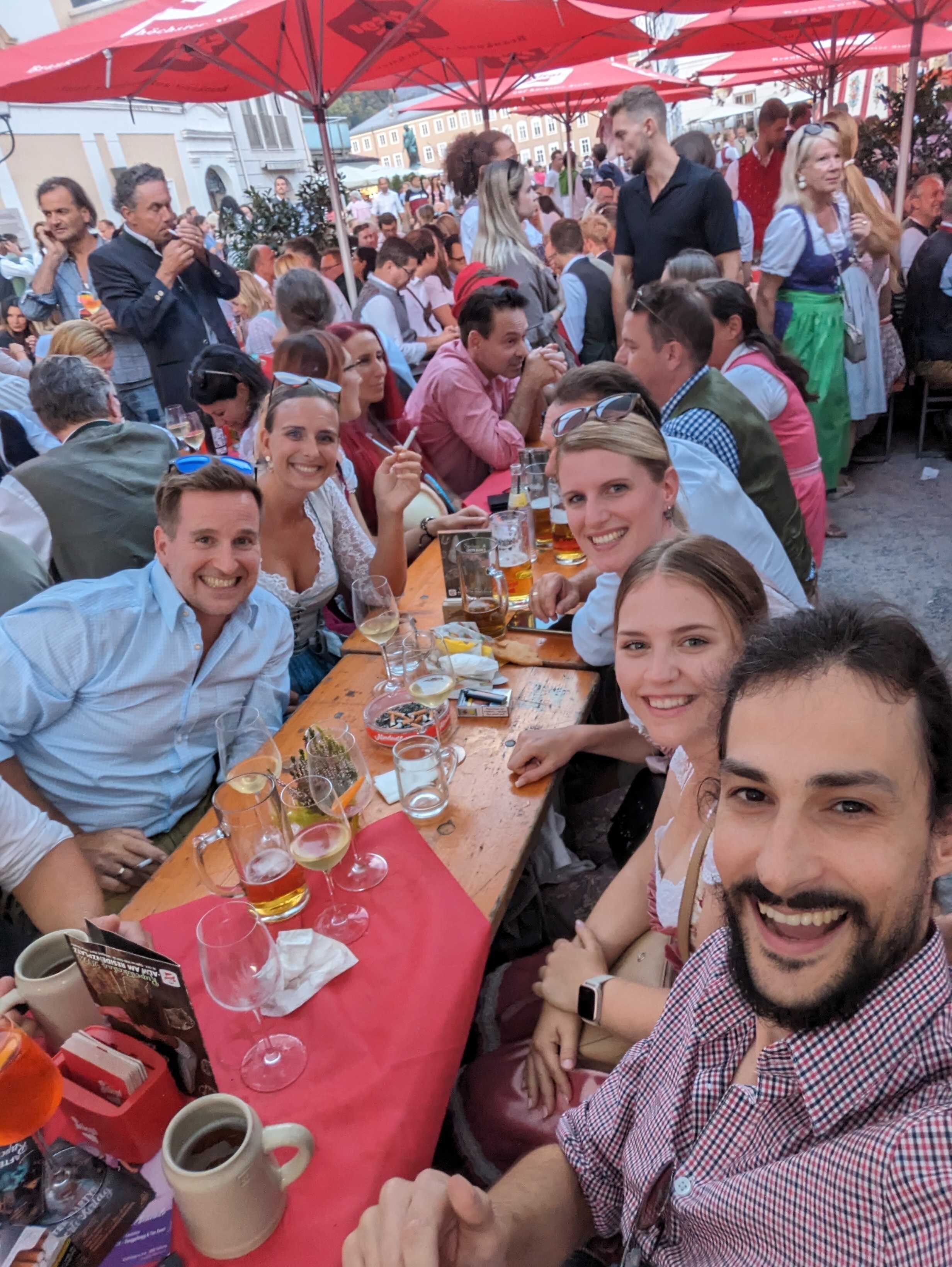 Eine Gruppe macht ein Selfie an einem vollen Biertisch im Gastgarten, in Tracht, mit Bierkrügen und Weingläsern unter roten Sonnenschirmen.