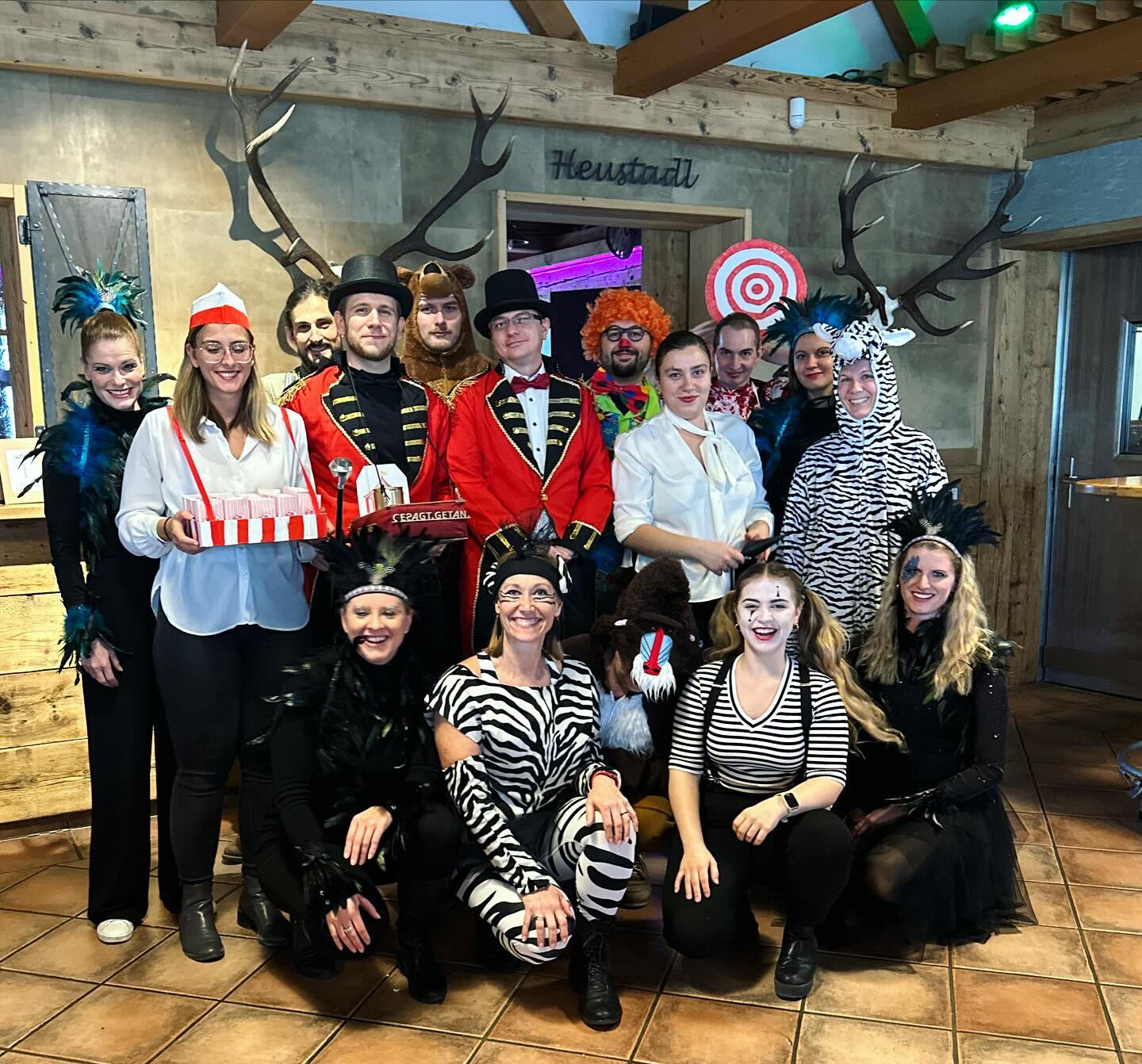 Verkleidete Gruppe im Heustadl: Tiere, Zirkusdirektoren und Clown posieren zum Faschings‑Gruppenfoto in einem rustikalen Gasthaus.