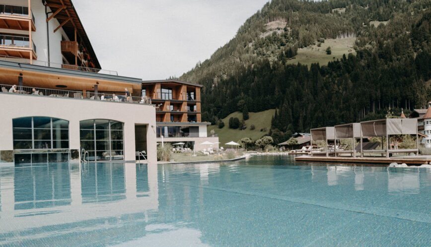 Infinity-Pool eines Alpenresorts mit Blick auf bewaldete Berghänge und Sonnendeck mit überdachten Liegen.