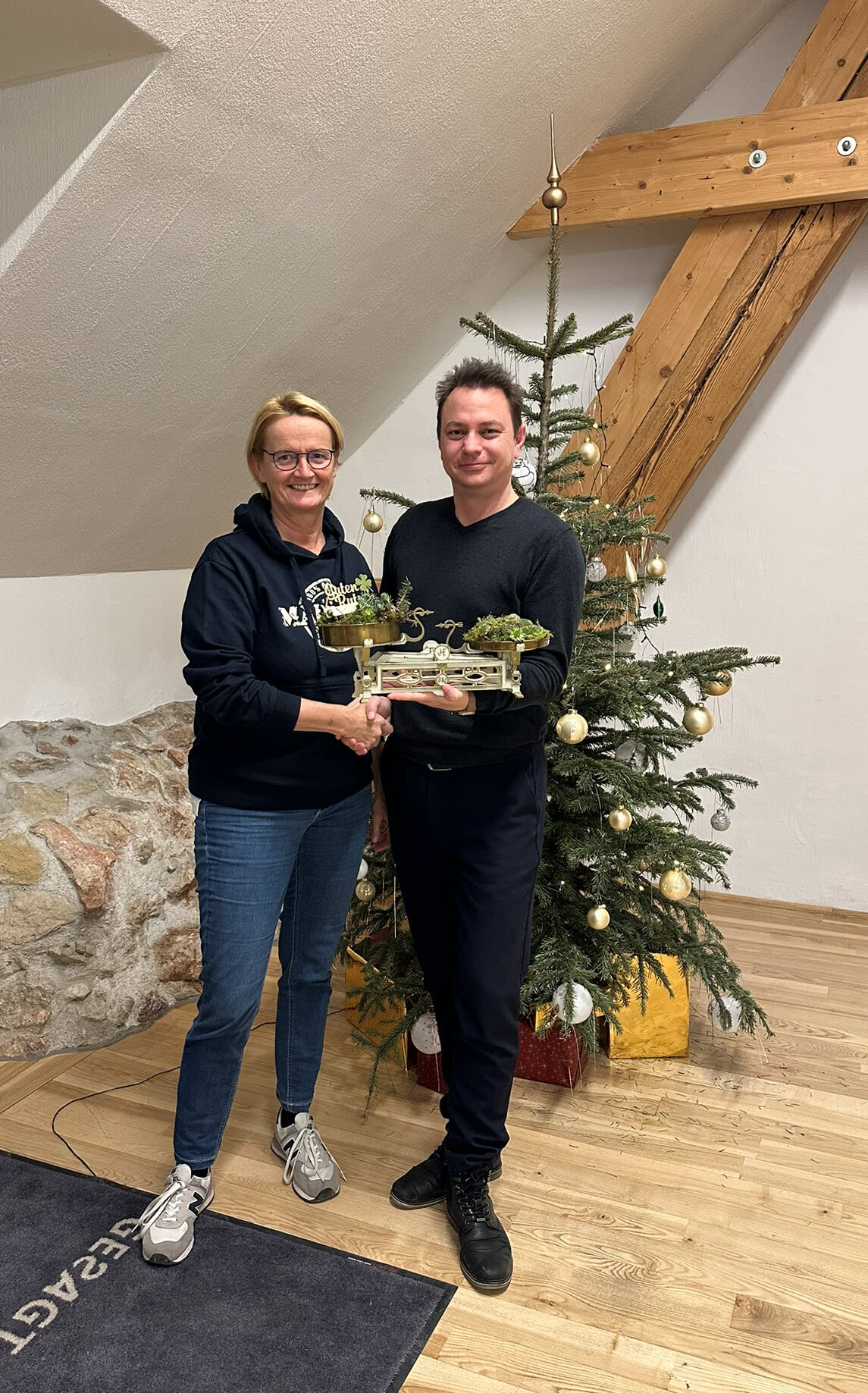 Zwei Personen reichen sich die Hand und halten eine alte Waage mit Pflanzen vor einem geschmückten Christbaum.