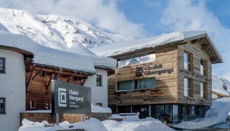 Chalet Obergurgl mit Holzfassade und Schild „Apartments & Suites“, vor tief verschneiten Ötztaler Alpen unter blauem Himmel.