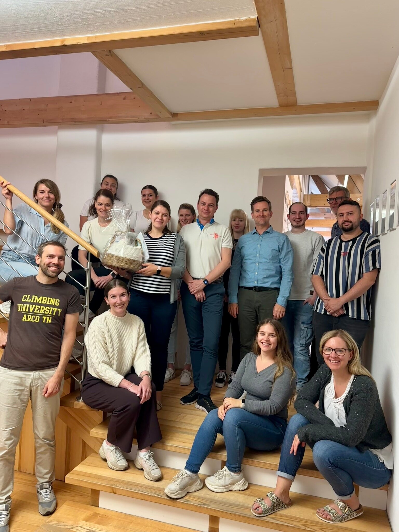 Teamfoto im Büro: Mitarbeitende sitzen und stehen auf einer Holztreppe, lächeln; eine Person hält einen Geschenkkorb.